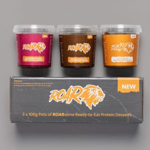 Trio of Roar protein desserts displayed against a grey background stacked on top of their box which is black with ROAR in orange writing with an orange lion head beside the logo