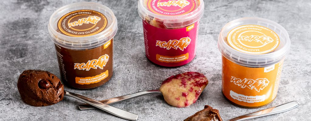 Three ROAR Protein pots chocolate, raspberry and jaffa displayed against a grey background with a spoonful of each beside