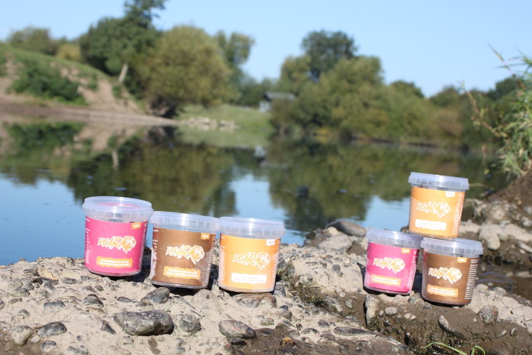 Six pots of ROAR protein of all three flavours placed on rocks beside a river with trees reflecting in the water and blue sky