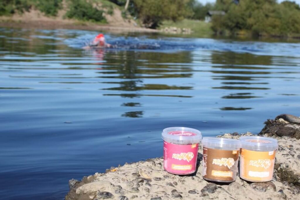 three ROAR protein pots rest beside a river with a swimmer in a red hat swimming towards them to eat after training as a triathlon nutrition hack