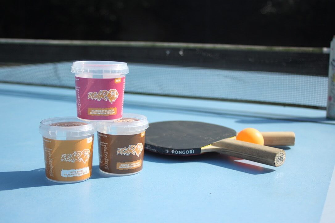 chocolate, jaffa and raspberry protein pots rest on a blue table tennis table with bats, ball and net beside