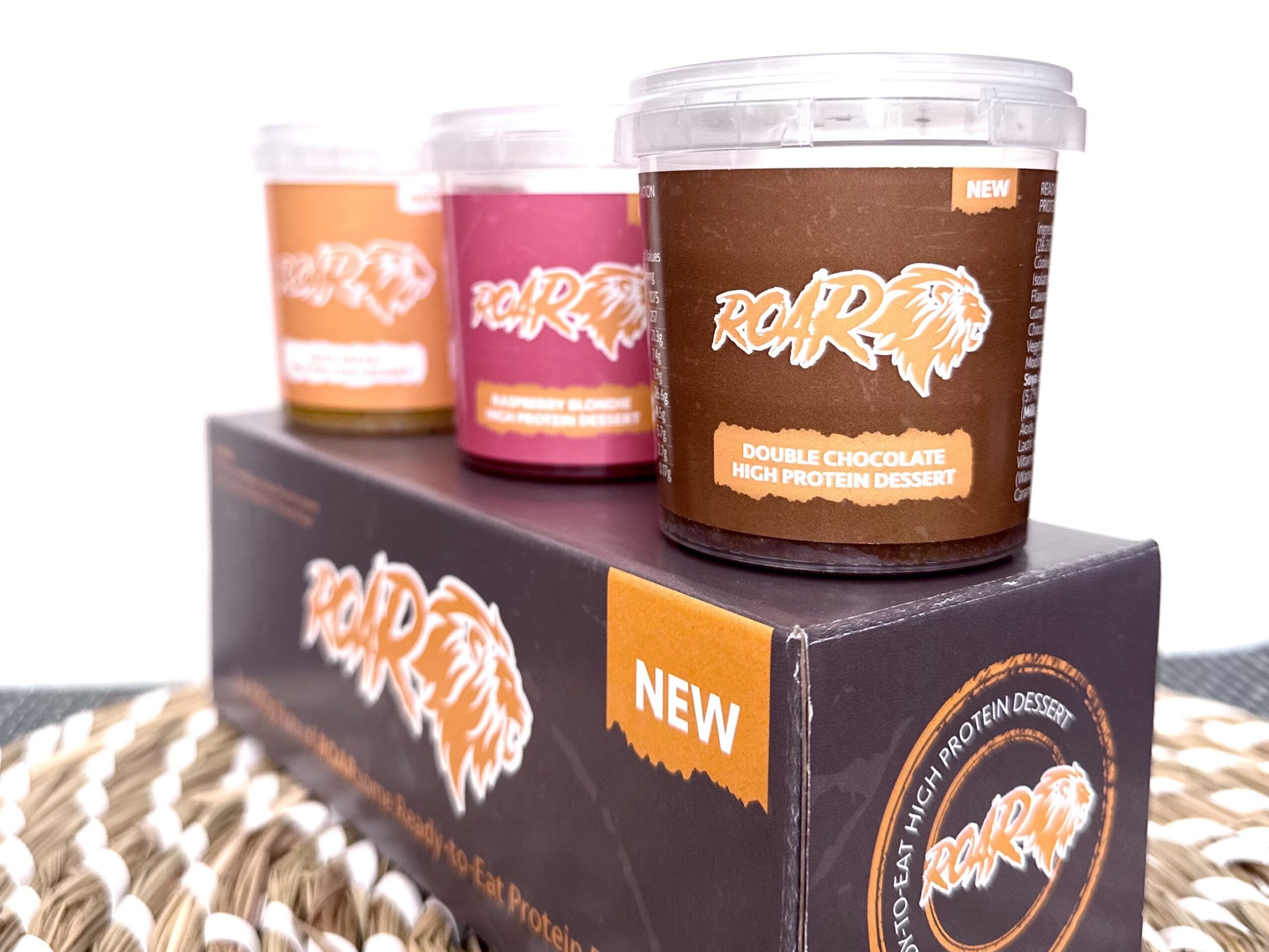 Three pots of ROAR protein dessert displayed on top of their box with the chocolate protein dessert at the front then raspberry then orange with a brown and white mat beneath and white background